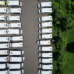 Vista aérea de uma frota de ônibus brancos alinhados em um pátio próximo a uma área verde.