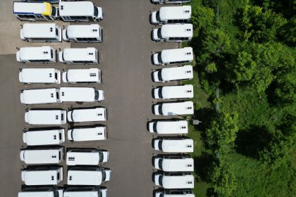 Vista aérea de uma frota de ônibus brancos alinhados em um pátio próximo a uma área verde.