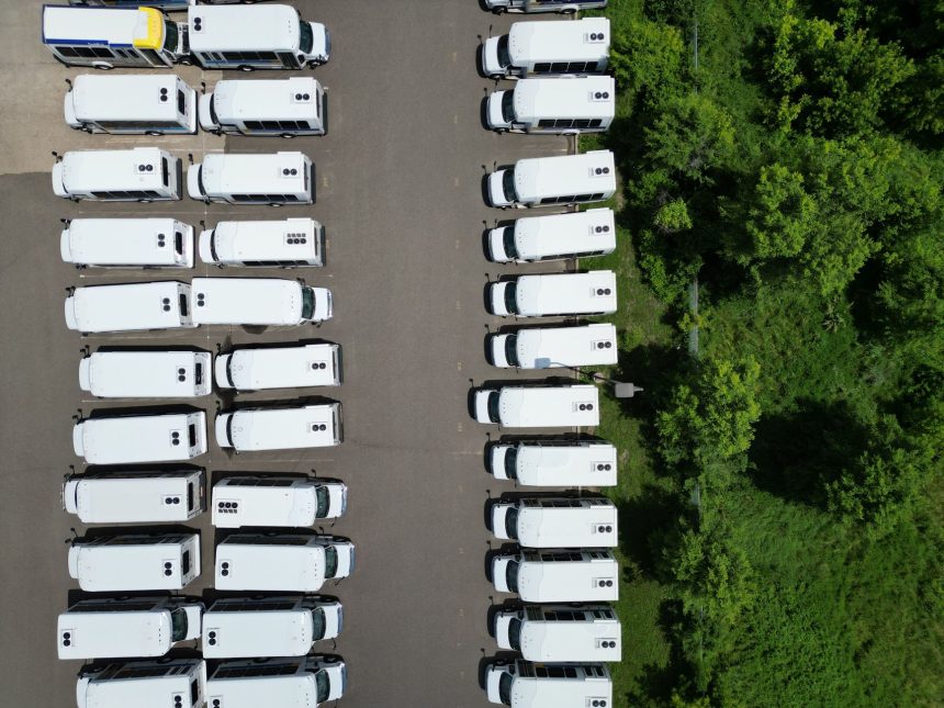 Vista aérea de uma frota de ônibus brancos alinhados em um pátio próximo a uma área verde.