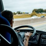 Motorista conduz ônibus em uma rodovia movimentada, com carros ao fundo.