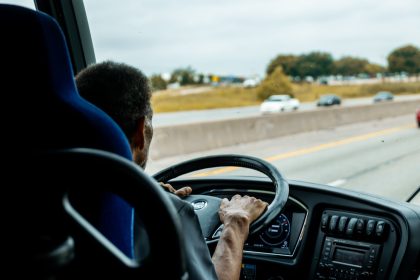 Motorista conduz ônibus em uma rodovia movimentada, com carros ao fundo.