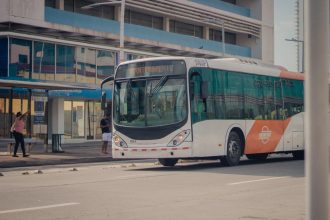 Passageiros embarcam em um ônibus urbano parado em um ponto de uma via movimentada.