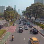 Via urbana movimentada em Jacarta, com carros, motocicletas e um ponto de ônibus ao lado da pista.