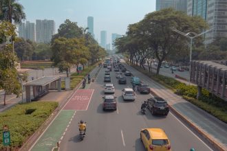 Via urbana movimentada em Jacarta, com carros, motocicletas e um ponto de ônibus ao lado da pista.